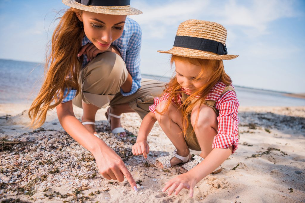 happy mother and daughter in straw hats collecting seashells at beach